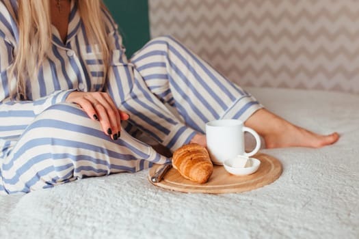 a woman in pajamas drinks coffee with a croissant on the bed . the concept of breakfast in bed.