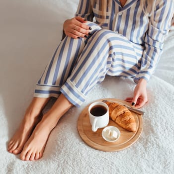 women's beautiful legs in pyjamas and next to a coffee with a croissant.the concept of coffee in bed . top view