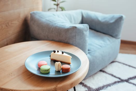 a cheese cake with blackcurrant and macarons on a turquoise plate sits on the edge of the coffee table. sweet breakfast concept