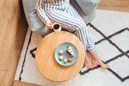 a woman in pajamas in the morning at a table drinks coffee and eats sweets cheesecake sweet macaroni . the concept of breakfast with sweets . top view