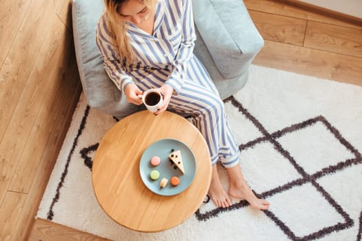 a woman in pajamas in the morning at a table drinks coffee and eats sweets cheesecake sweet macaroni . the concept of breakfast with sweets . top view