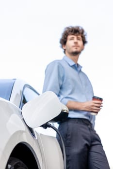 Progressive eco-friendly concept of focus parking EV car at public electric-powered charging station in city with blur background of businessman leaning on recharging-electric vehicle with coffee.