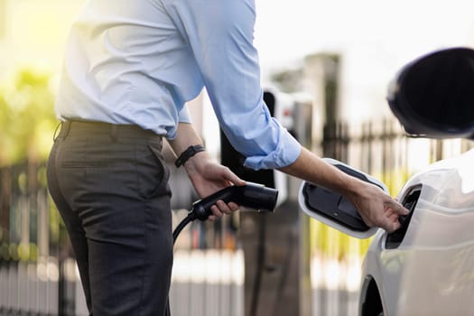 Closeup progressive man holding EV charger plug from public charging station for electric vehicle with background of residential building as concept eco-friendly sustainability energy car concept.