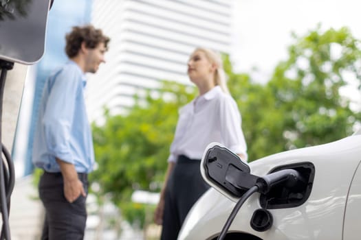 Focus parking-electric car connected to public charging station with blur progressive businesspeople holding coffee, residential building apartment and condo background for eco-friendly concept.