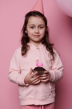 Vertical advertising studio shot on pink color background, lovely little child girl in pink party hat with a birthday cake in her hands, cutely smiling looking at camera. Happy Childhood concept