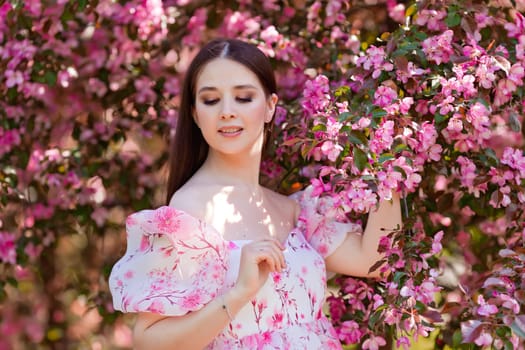 A brunette in a pink dress standing near pink blooming apple trees, holding a blooming branch, in the spring in the garden, looks to the down. Copy space