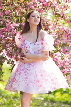 One brunette girl in light pink dress, is standing near a pink blooming apple trees, in the spring in the garden. Looks to the side. Vertical. Copy space