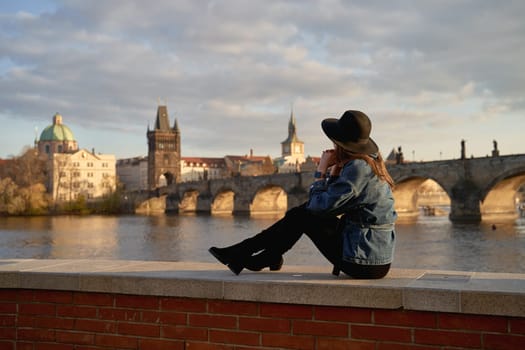 Stylish young beautiful woman earing black hat in Prague with Charles Bridge on background. Elegant retro lady fine art portrait.