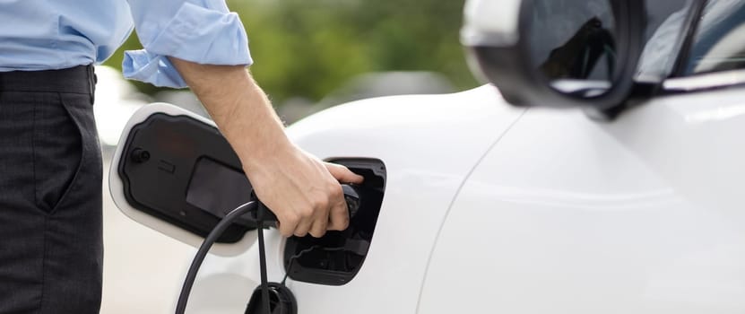 Closeup progressive man holding EV charger plug from public charging station for electric vehicle with background of residential building as concept eco-friendly sustainability energy car concept.