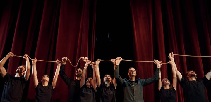 Holding rope in hands above the heads. Group of actors in dark colored clothes on rehearsal in the theater.