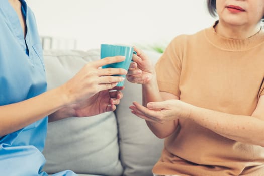 Female care taker serving her contented senior patient with a cup of coffee at home, smiling to each other. Medical care for pensioners, Home health care service.