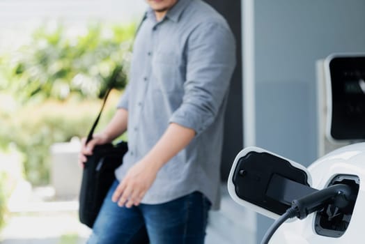 Focus electric car charging at home charging station with blurred progressive man walking in the background. Electric car using renewable clean for eco-friendly concept.