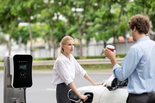 Progressive businessman and businesswoman with electric car parking and connected to public charging station before driving around city center. Eco friendly rechargeable car powered by clean energy.