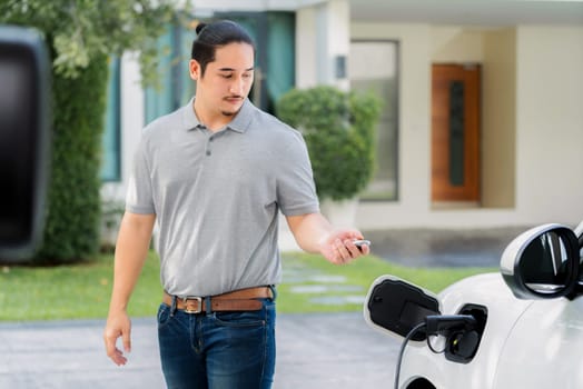 Progressive asian man install cable plug to his electric car with home charging station in the backyard. Concept use of electric vehicles in a progressive lifestyle contributes to clean environment.