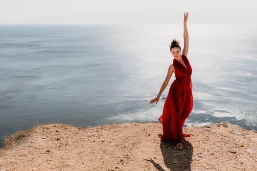 Side view a Young beautiful sensual woman in a red long dress posing on a rock high above the sea during sunrise. Girl on the nature on blue sky background. Fashion photo.