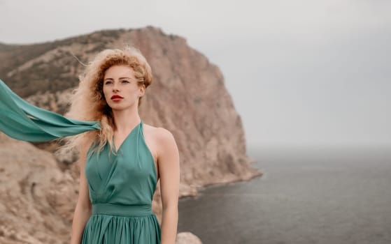 Side view a Young beautiful sensual woman in a mint long dress posing on a volcanic rock high above the sea during sunset. Girl on the nature on overcast sky background. Fashion photo