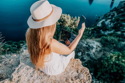 Successful business woman in yellow hat working on laptop by the sea. Pretty lady typing on computer at summer day outdoors. Freelance, travel and holidays concept.