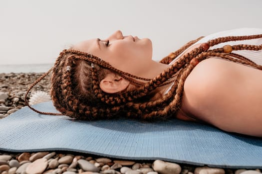 Woman yoga sea. Well looking middle aged woman with braids dreadlocks in white leggings and tops doing stretching pilates on yoga mat near sea. Female fitness yoga routine concept. Healthy lifestyle