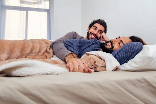 gay male couple relaxing lying on bed with clasped hands, concept of real people lifestyle and love between people of the same sex