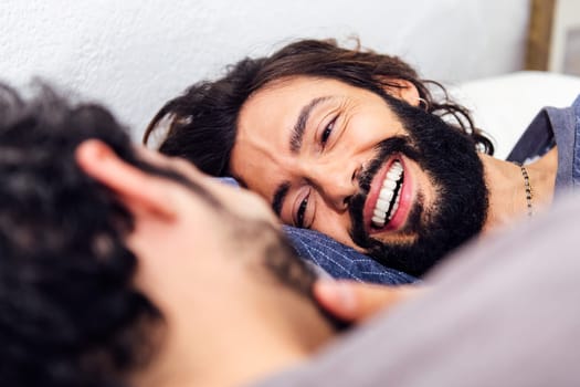 smiling happy man relaxed in bed stroking his boyfriend's face, concept of real people lifestyle at home and love between people of the same sex