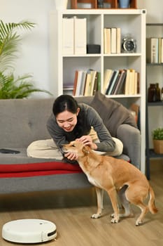 Young woman and dog sitting in living room with robot vacuum cleaner cleaning the floor. Smart home and automatic cleaning concept.