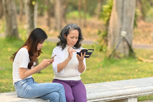 Image of middle age woman and daughter using mobile phone while sitting on a bench in calm nature park.