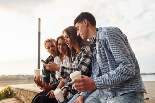 Sunny weather. Drinking coffee. Group of young cheerful friends that is outdoors having fun together.