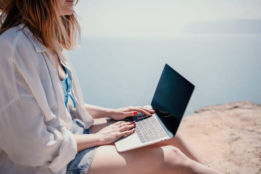 Successful business woman in yellow hat working on laptop by the sea. Pretty lady typing on computer at summer day outdoors. Freelance, travel and holidays concept.