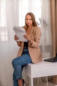 professional woman sits at desk in home office, positive female lawyer reads documents. She is wearing a beige jacket and jeans