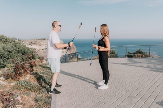 Happy Middle aged couple or friends practicing nordic walking in park near sea. Mature couple with trekking poles walking, practicing Nordic walking outdoors. Aging youthfully and sport concept.