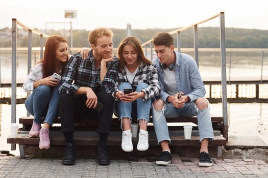 Sitting near lake and embracing each other. Group of young cheerful friends that is outdoors having fun together.