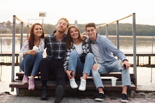 Sitting near lake and embracing each other. Group of young cheerful friends that is outdoors having fun together.