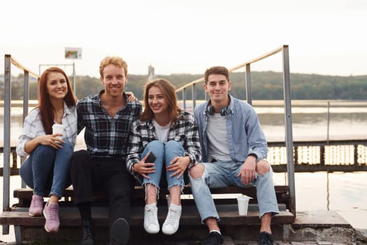 Sitting near lake and embracing each other. Group of young cheerful friends that is outdoors having fun together.