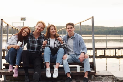 Sitting near lake and embracing each other. Group of young cheerful friends that is outdoors having fun together.