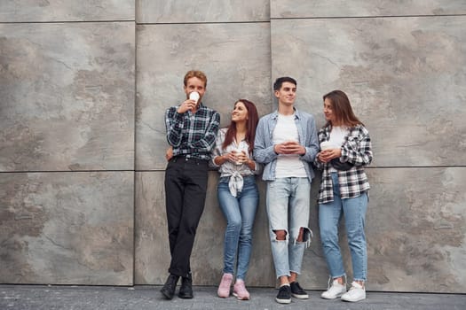 Group of young positive friends in casual clothes standing together against grey wall with cups of drink in hands.