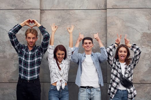 Making different gestures. Group of young positive friends in casual clothes standing together against grey wall.