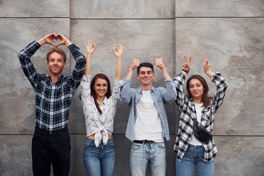 Making different gestures. Group of young positive friends in casual clothes standing together against grey wall.