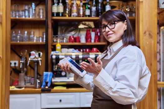 Woman in apron worker manager of bar restaurant cafe holding wireless modern banuov payment terminal for processing credit card payments. Cashless technology, money service, work small business staff