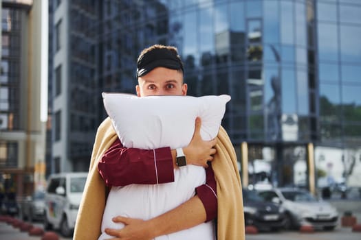 Embracing the pillow. Young man in pajama is outdoors on the street. Feels sleepy.