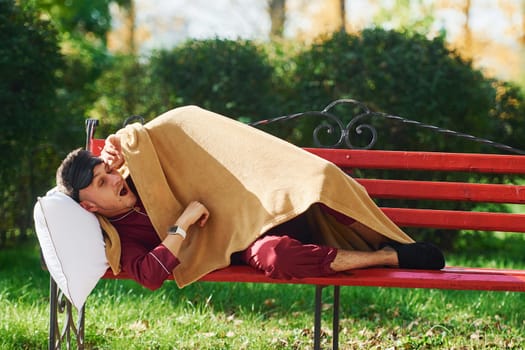 Lying down on bench in park. Young man in pajama is outdoors on the street. Feels sleepy.