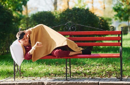 Lying down on bench in park. Young man in pajama is outdoors on the street. Feels sleepy.