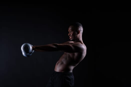 Confident young man shirtless portrait training with dumb-bell against black background.