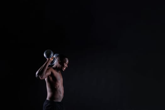 Confident young man shirtless portrait training with dumb-bell against black background.