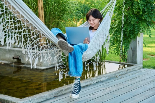 Teenage girl relaxing in hammock using laptop for leisure study. Adolescence, students, high school, technology, lifestyle, youth concept