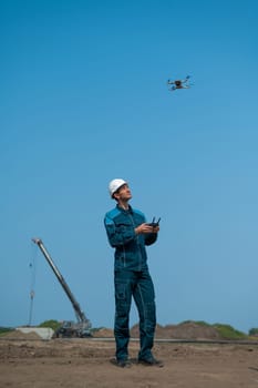 A man in a helmet and overalls controls a drone at a construction site. The builder carries out technical oversight