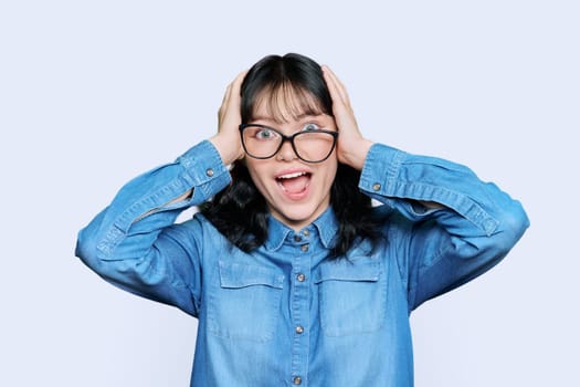 Shocked anxious young woman in panic holding her hands behind her head with her mouth open on white studio background. Teenage student with glasses in hysterics, shock stress helplessness anxiety