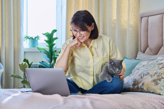 Middle aged woman at home on couch with laptop and cat. Mature 40s female looking at screen, on bed with pet. Work at home, technology, leisure, freelancing, lifestyle, people and animals concept