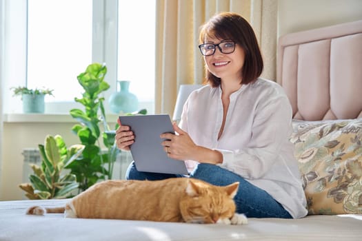 Middle aged woman at home on couch with laptop and cat. Mature 40s female looking at camera, on bed with pet. Work at home, technology, leisure, freelancing, lifestyle, people and animals concept