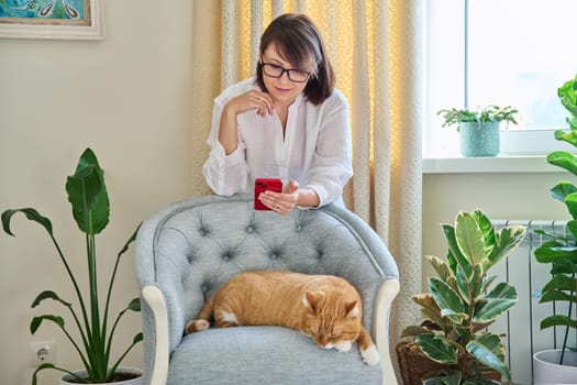 Middle aged woman using smartphone, in home interior with sleeping cat on armchair, houseplants. Lifestyle, comfort, green pets people animals concept