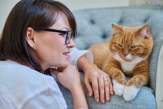 Middle aged woman talking with ginger pet cat, home interior background. Friendship, love, care, leisure, lifestyle, animals people house concept